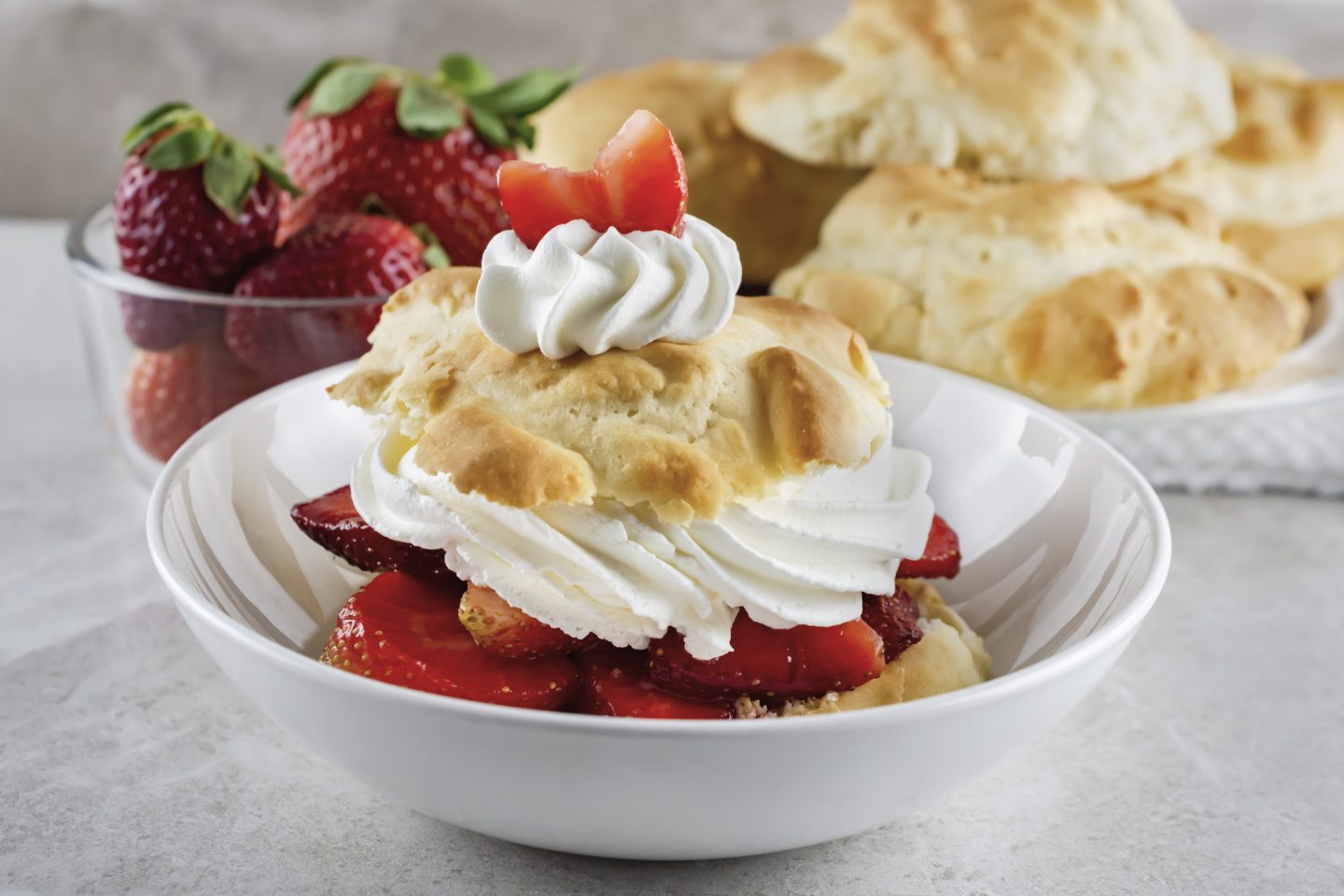 Strawberry shortcake in shallow white bowl with whipped cream and garnished with a sliced strawberry. Bowl of strawberries in background with a plate of pastries.