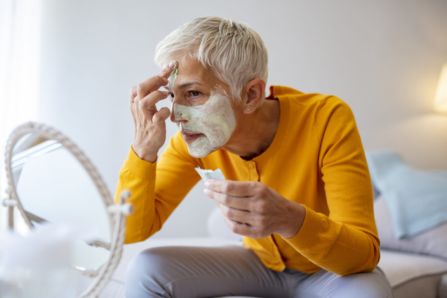Senior woman with a facial mask. Smiling mature woman applying rejuvenating mask on her face skin. Elderly female enjoy making face massage, peeling, put on mask.