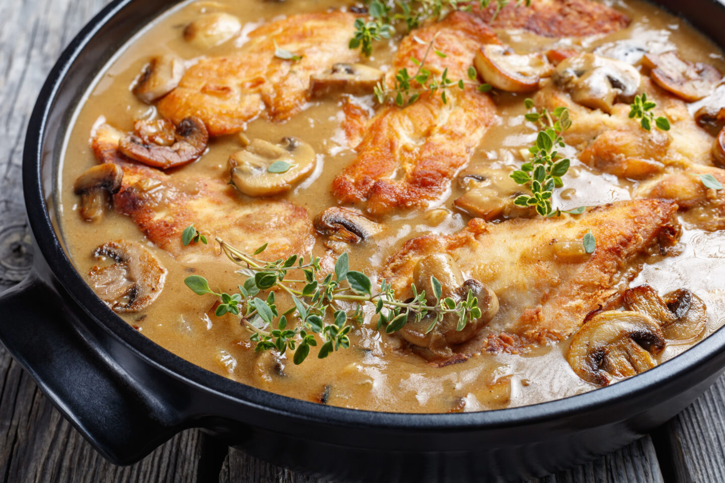 golden pan-fried chicken cutlets and mushrooms in a rich Marsala wine sauce in a black ceramic dish on a wooden table, horizontal view from above