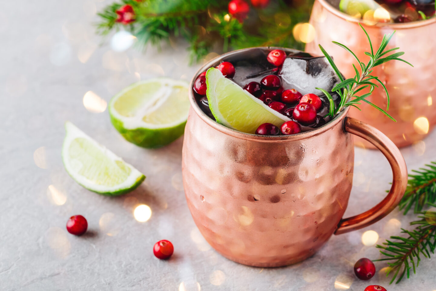 Christmas Moscow Mule ice-cold Cocktail in a copper cup with cranberries, lime, and rosemary on gray stone background.