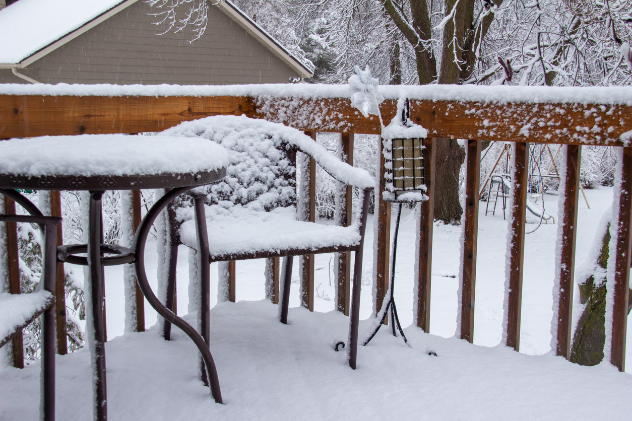 This image shows a close up view of an outdoor wooden deck with a round table and chairs, covered with thick snow during a winter blizzard.