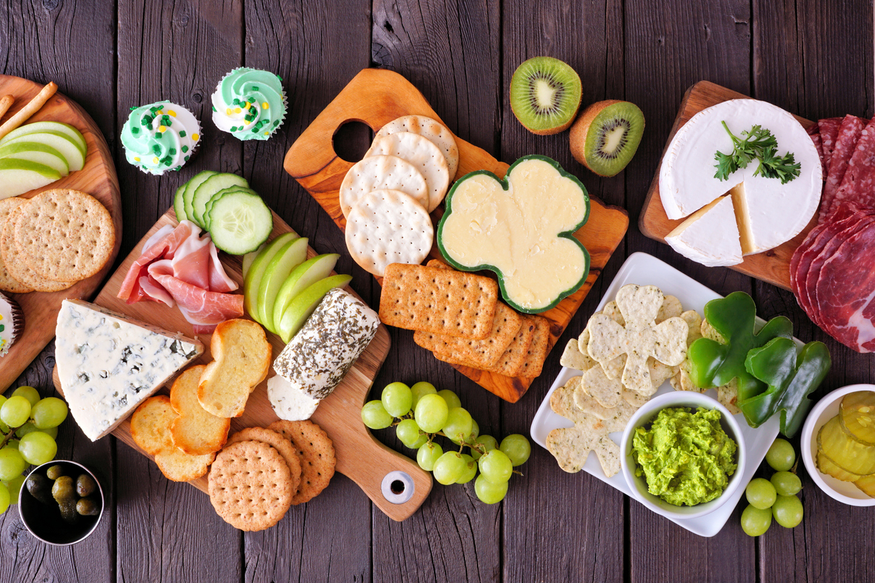 St Patricks Day theme charcuterie table scene against a wood background. Selection of cheese, meat, fruit and vegetable appetizers.