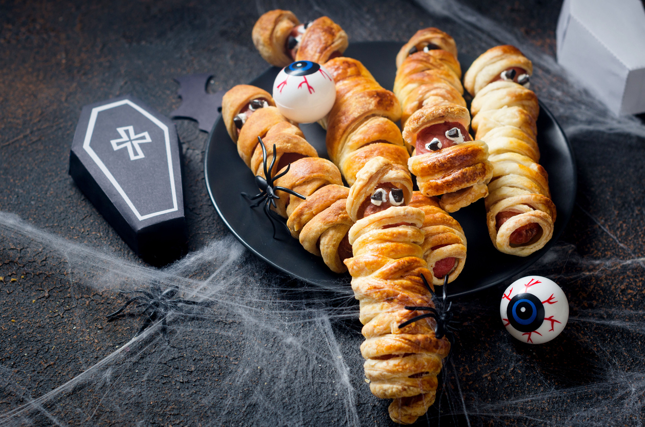 Spooky sausage mummies in dough in black plate with graves on dark table. Halloween funny food idea for kids party