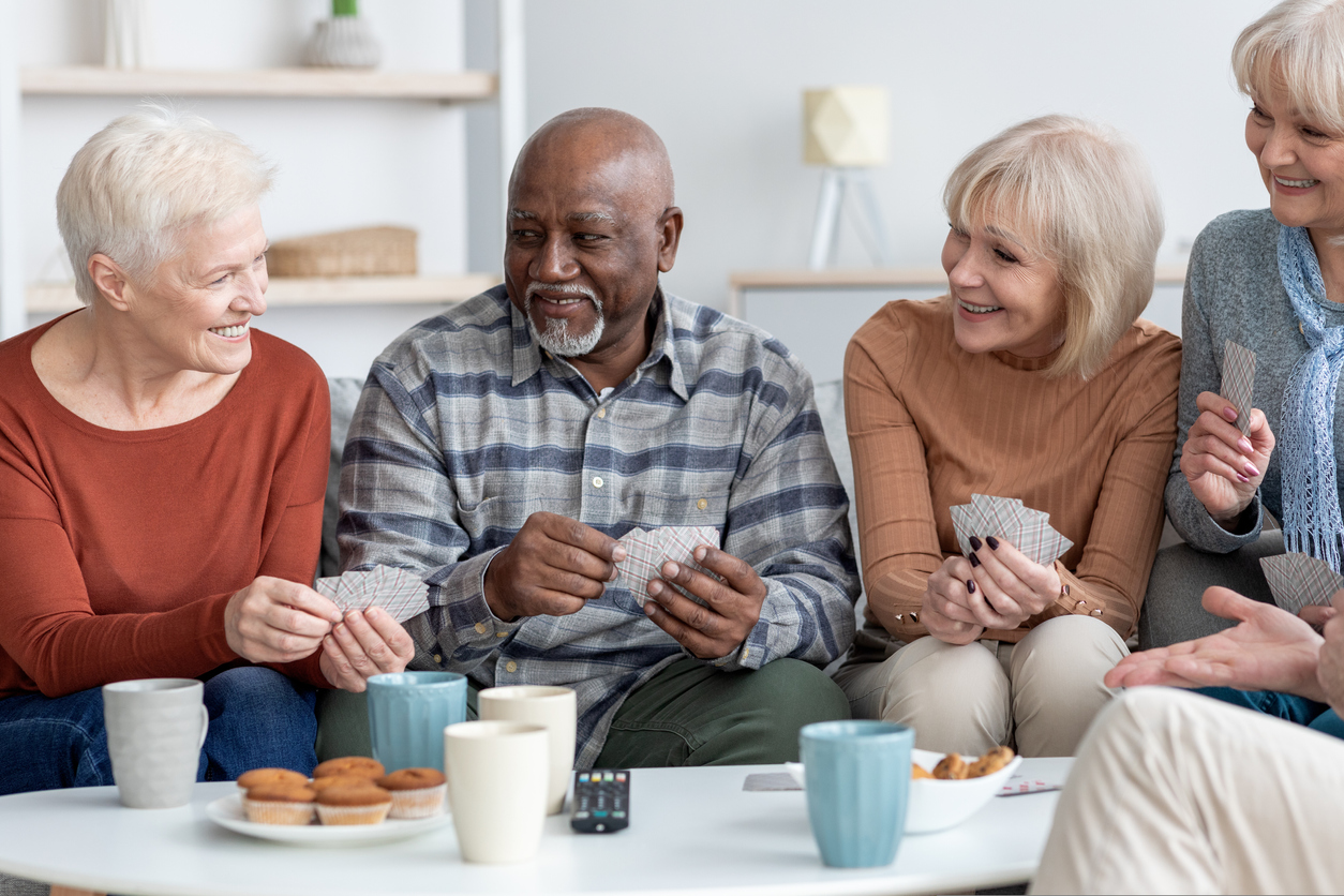 International group of cheerful elderly people men and women in casual outfits chilling together at sanatorium, sitting on sofa in living room, playing card games, drinking tea, chatting