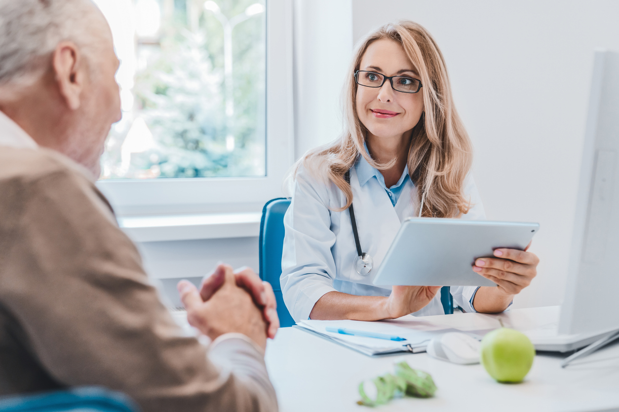 Middle aged female doctor nutritionist in white medical coat consulting her old patient using digital tablet