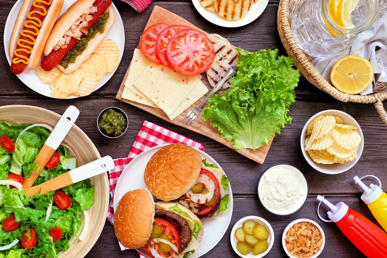Summer BBQ or picnic table scene with hamburgers, hotdogs, salad and snacks. Overhead view on a dark wood background.
